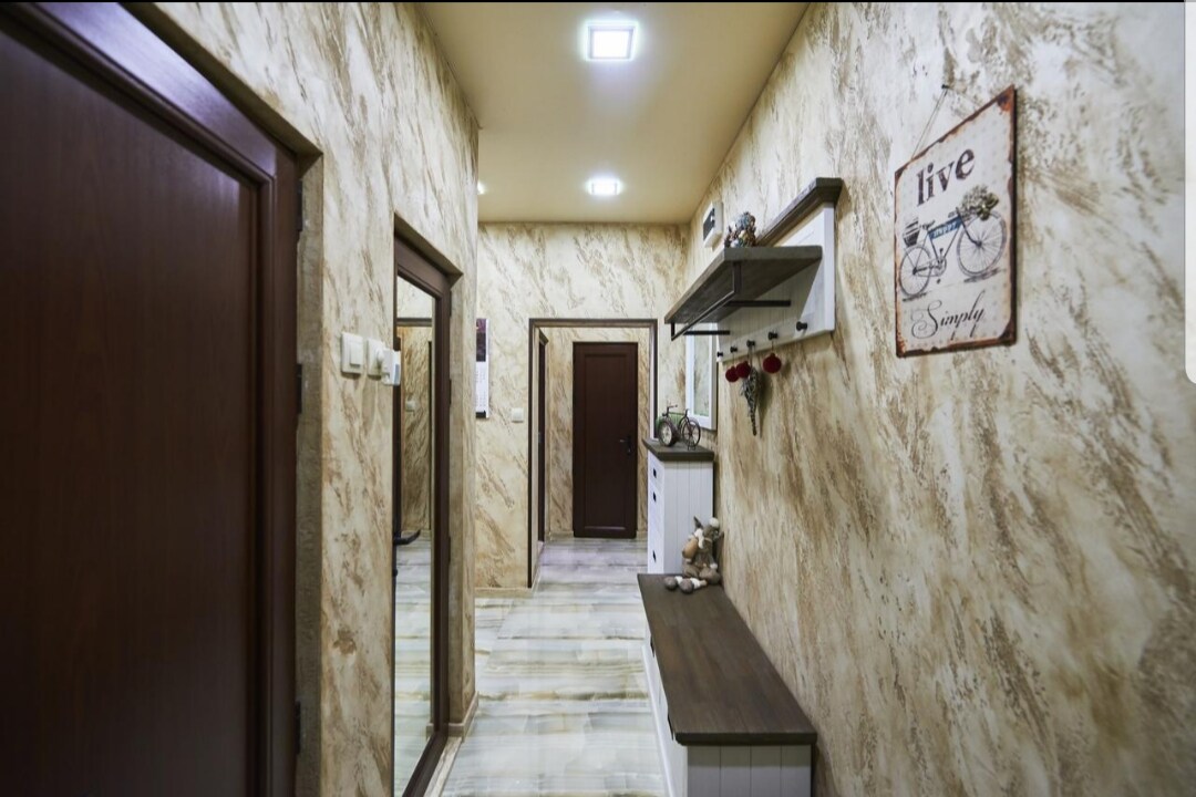A well-lit hallway features intricate textured walls in neutral tones. A narrow console table is positioned against one wall, accompanied by decorative items. A full-length mirror enhances the space, while a door at the end leads to another room.
