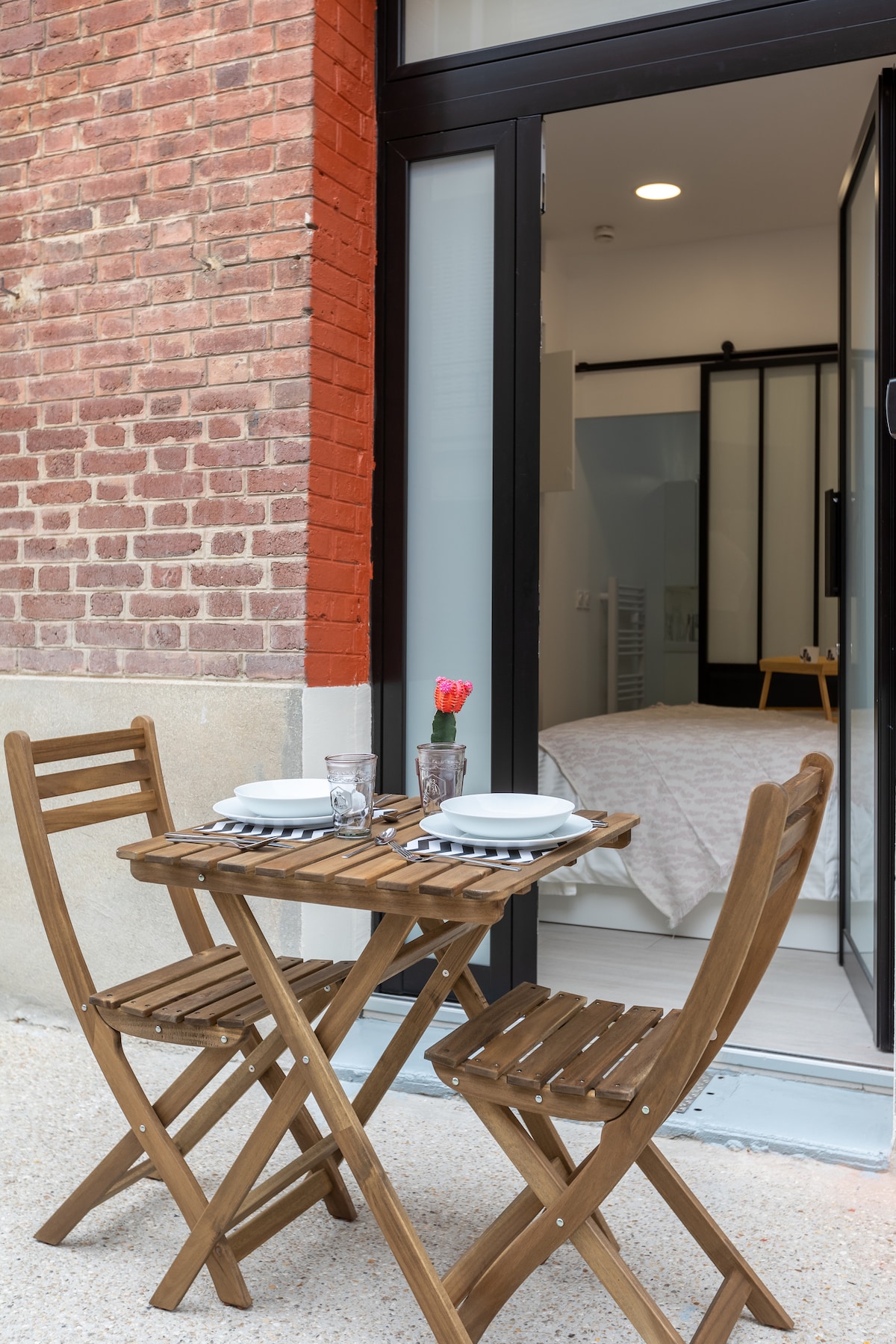 A small, wooden dining table is set on a private patio, accompanied by two folding chairs. A flower in a vase adds a touch of color to the neatly arranged plates and glasses. The patio area is accessible through glass doors, showcasing a light-filled interior.