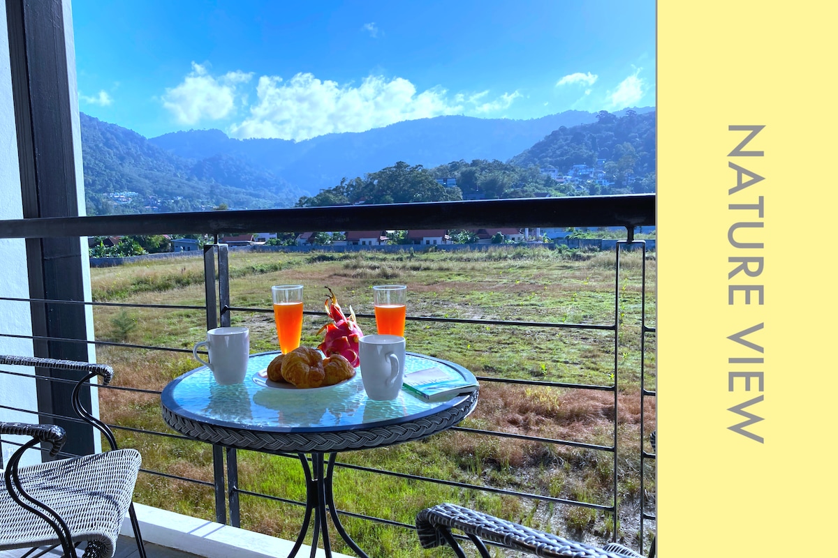 A balcony table is set with two glasses of orange juice and a white cup, accompanied by a croissant and a colorful fruit selection. In the background, a lush green landscape is visible, framed by rolling hills and an expansive blue sky.