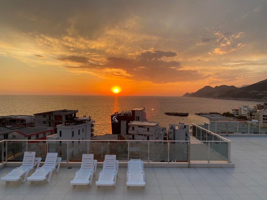 A spacious terrace is featured with several white lounge chairs, facing a breathtaking sunset over the ocean. The sky is painted in warm hues of orange and pink, while buildings line the horizon against the tranquil sea.