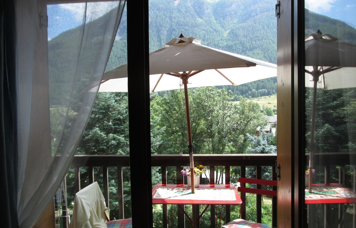A balcony is framed by large sliding doors, revealing an outdoor dining setup with a table and an umbrella. Lush trees and scenic mountains are visible in the background, contributing to a serene and natural environment.