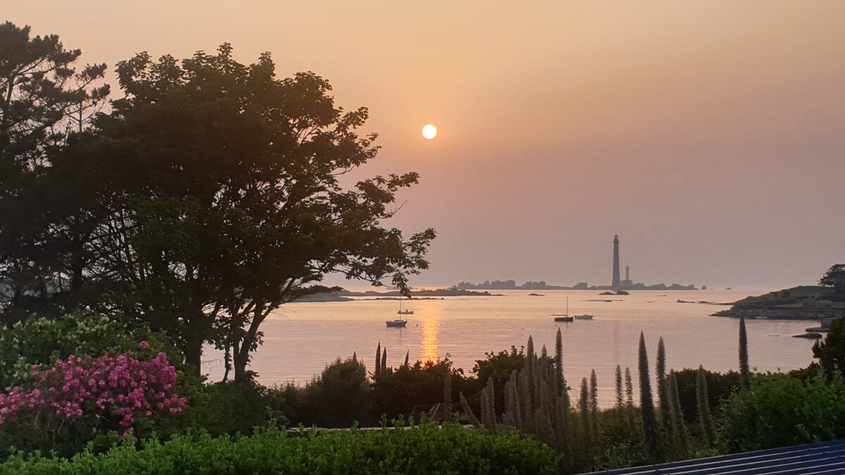 A serene seascape is captured during sunset, with the sun setting over calm waters. The silhouette of the Isle Vierge lighthouse stands tall in the distance, framed by trees and blooming flowers in the foreground.