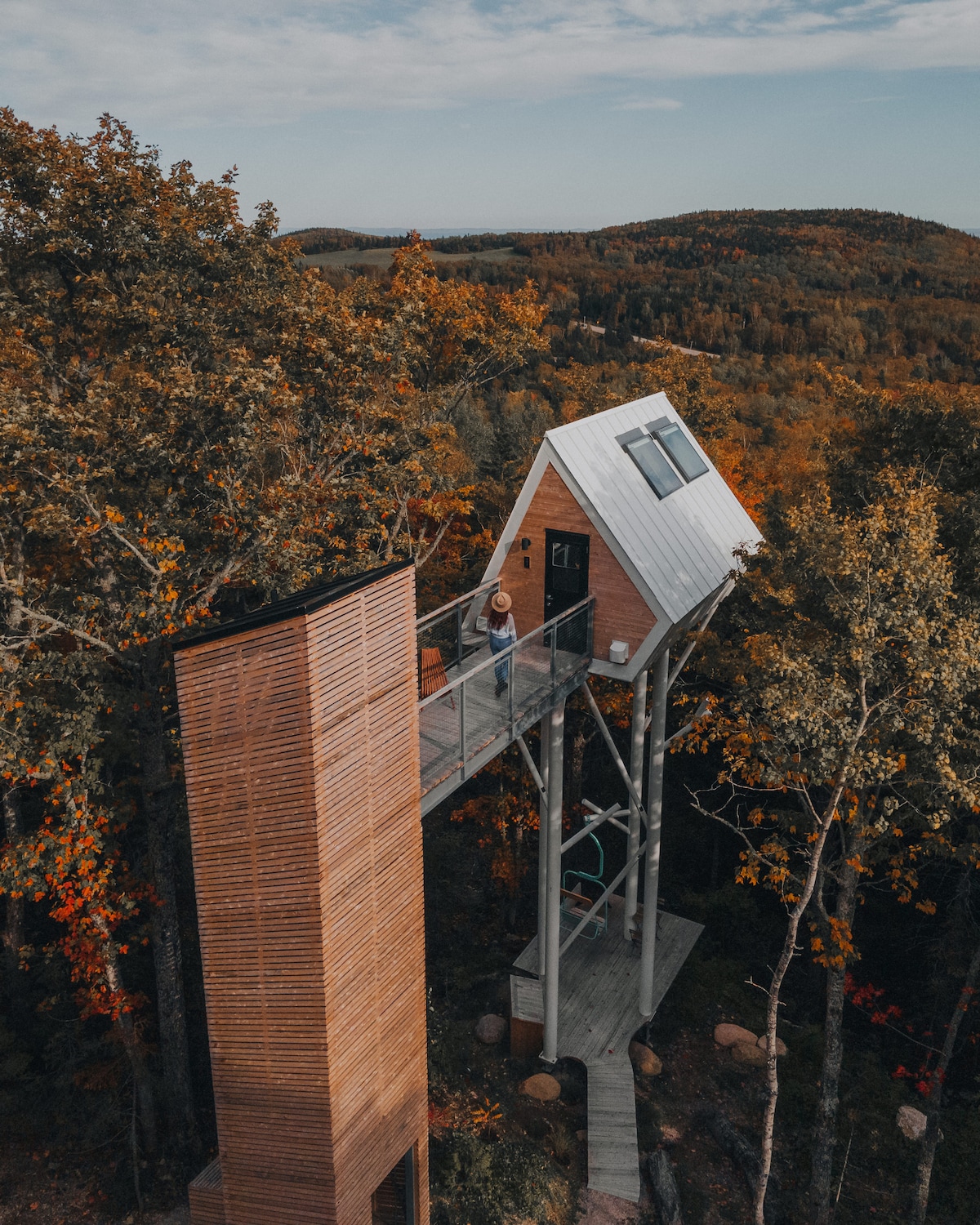 A modern cabin is elevated among autumn-colored trees, showcasing a unique architectural design with a sloped roof. A wooden walkway connects to the entrance, surrounded by natural scenery. The overall setting highlights the serene beauty of the landscape.