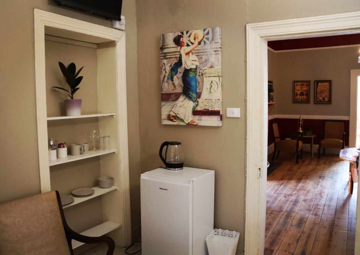 A corner of a shared living area is depicted, featuring a small refrigerator beside a kettle. A shelf holds glassware and dishes, while a potted plant adds a touch of greenery. An art piece hangs on the wall, enhancing the space with color.