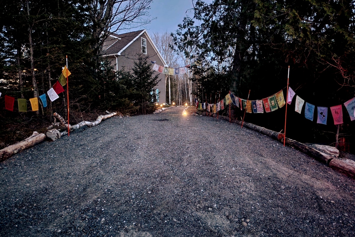 A gravel driveway is flanked by trees, with colorful prayer flags suspended between posts. Soft lighting guides the path to the welcoming entrance of a home nestled in a serene forested setting.