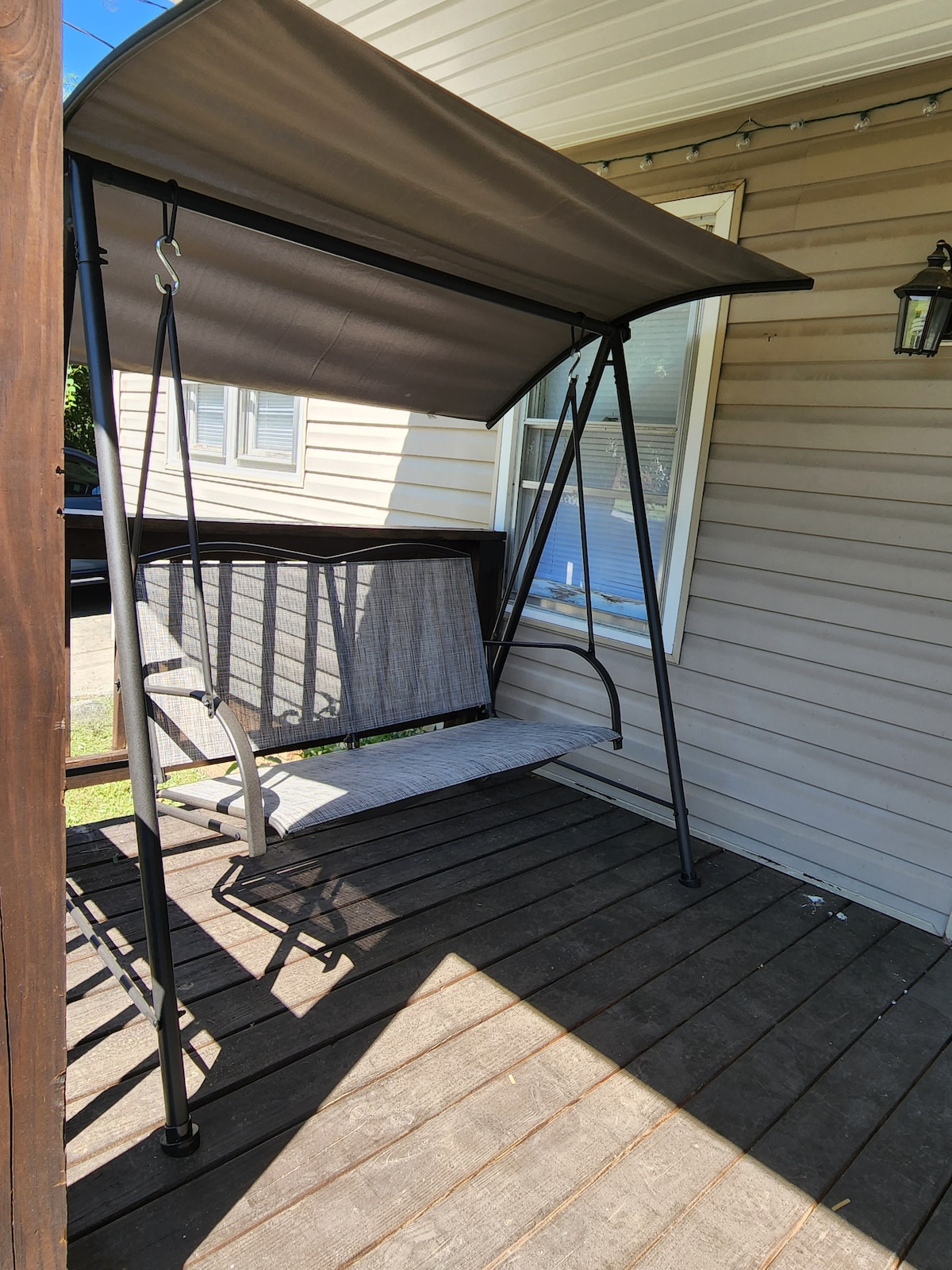 A covered outdoor swing is positioned on a wooden porch, providing shaded seating. The swing features a sturdy metal frame and a comfortable seat, with light filtering through a nearby window. The floorboards are made of dark wood, creating a cozy outdoor space.