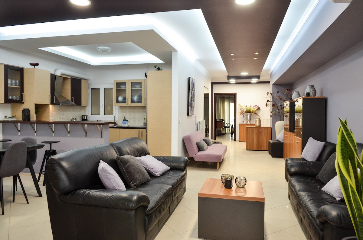 A spacious living area is highlighted by a mix of leather sofas, complemented by purple accent pillows. A wooden coffee table sits centrally, surrounded by bar stools. The open layout connects to a modern kitchen area with light cabinetry, illuminated by soft ceiling lighting.