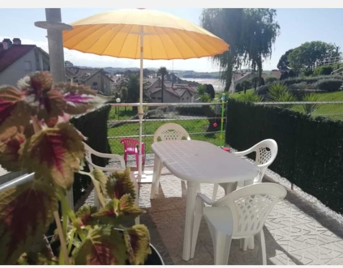 A terrace area is visible featuring a white outdoor table surrounded by four plastic chairs. A bright yellow umbrella provides shade over the table. Lush greenery and a distant view of the water create a serene outdoor setting.