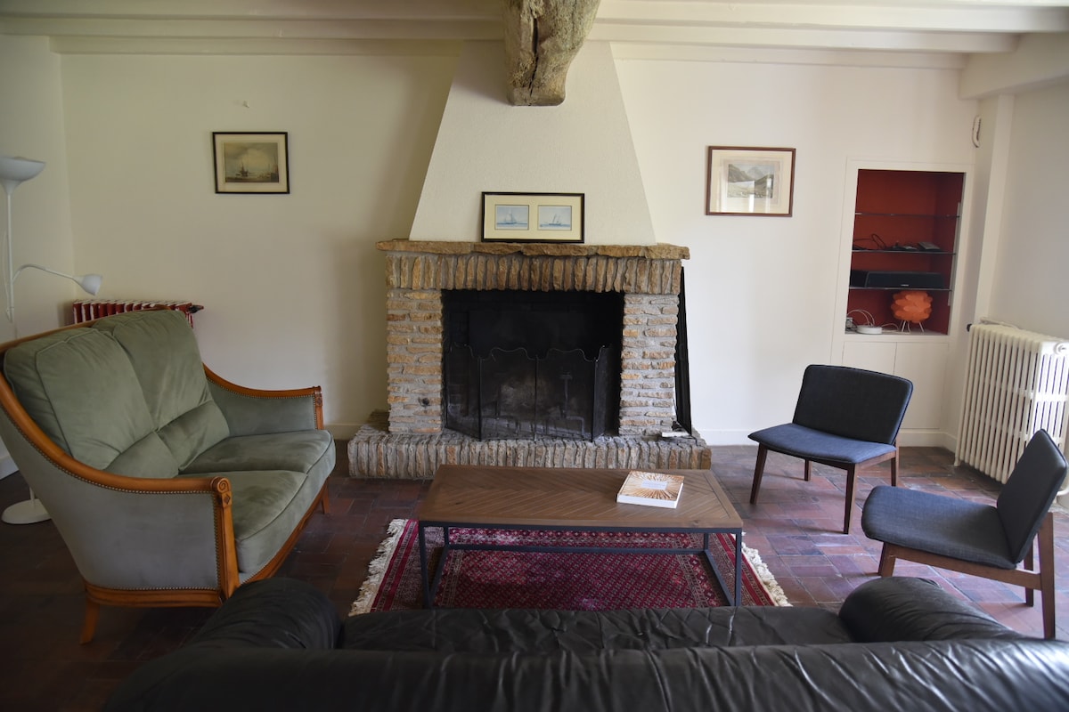 A comfortable living area features a traditional stone fireplace as a focal point. Two upholstered armchairs and a cozy sofa complement a wooden coffee table, set over a patterned rug. Soft natural light illuminates the space, highlighting the simple decor and exposed beam ceiling.