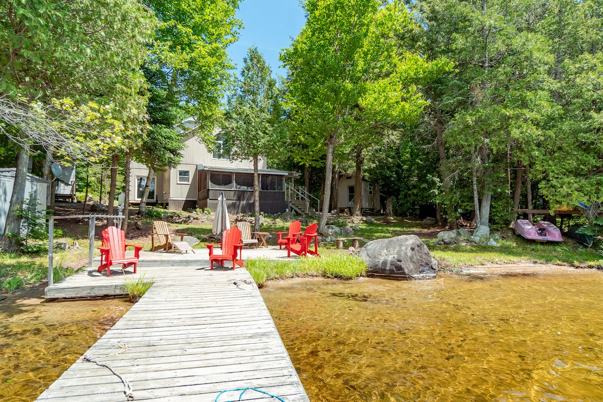A wooden dock extends over a sandy shoreline, with a gentle gradient into clear water. Muskoka chairs in red are positioned on the dock, surrounded by tall trees and grassy areas. The cottage is visible in the background, providing a harmonious blend with nature.