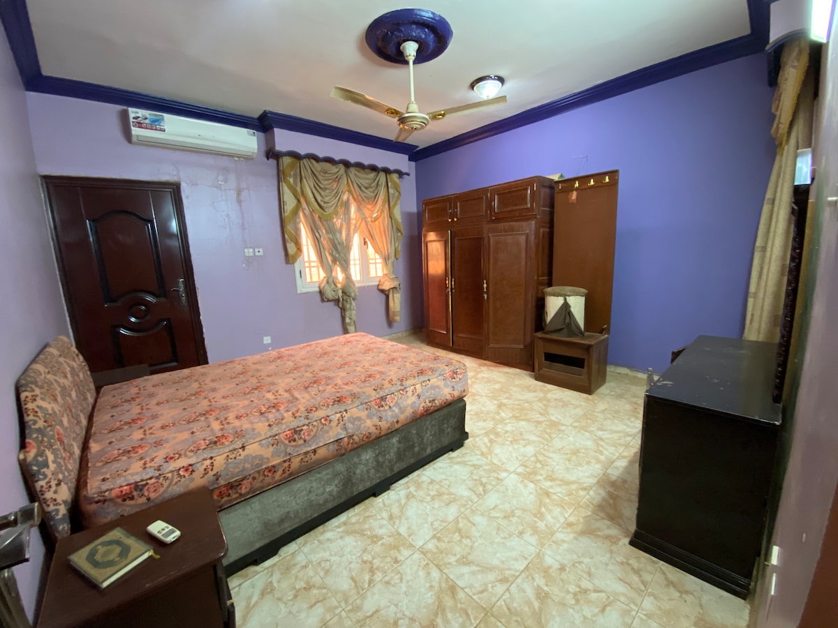 A spacious bedroom features a floral-patterned bedspread atop a large bed. Elegant drapes frame the window, while a wooden wardrobe and a chest of drawers provide storage. Neutral tiles complement the room's purple walls, and an air conditioning unit is mounted above.