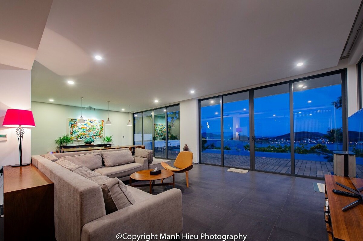 A spacious living area features a comfortable sofa and an accent chair, with large glass doors that lead to a terrace. The room is softly illuminated, highlighting a colorful artwork on the wall and offering expansive views of the surrounding landscape.