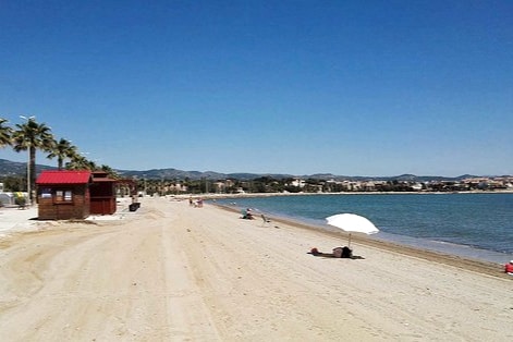 A sandy beach is visible, stretching along a clear blue shoreline. Palm trees line the beach, with a wooden beach hut situated nearby. A few beachgoers can be seen relaxing under an umbrella, while distant hills create a peaceful backdrop.