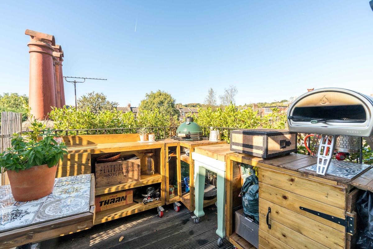 An outdoor kitchen area is shown, featuring wooden countertops, a small grill, and various potted plants. The space offers views of greenery and a clear blue sky, with a chimney stack visible in the background, contributing to a relaxed outdoor atmosphere.
