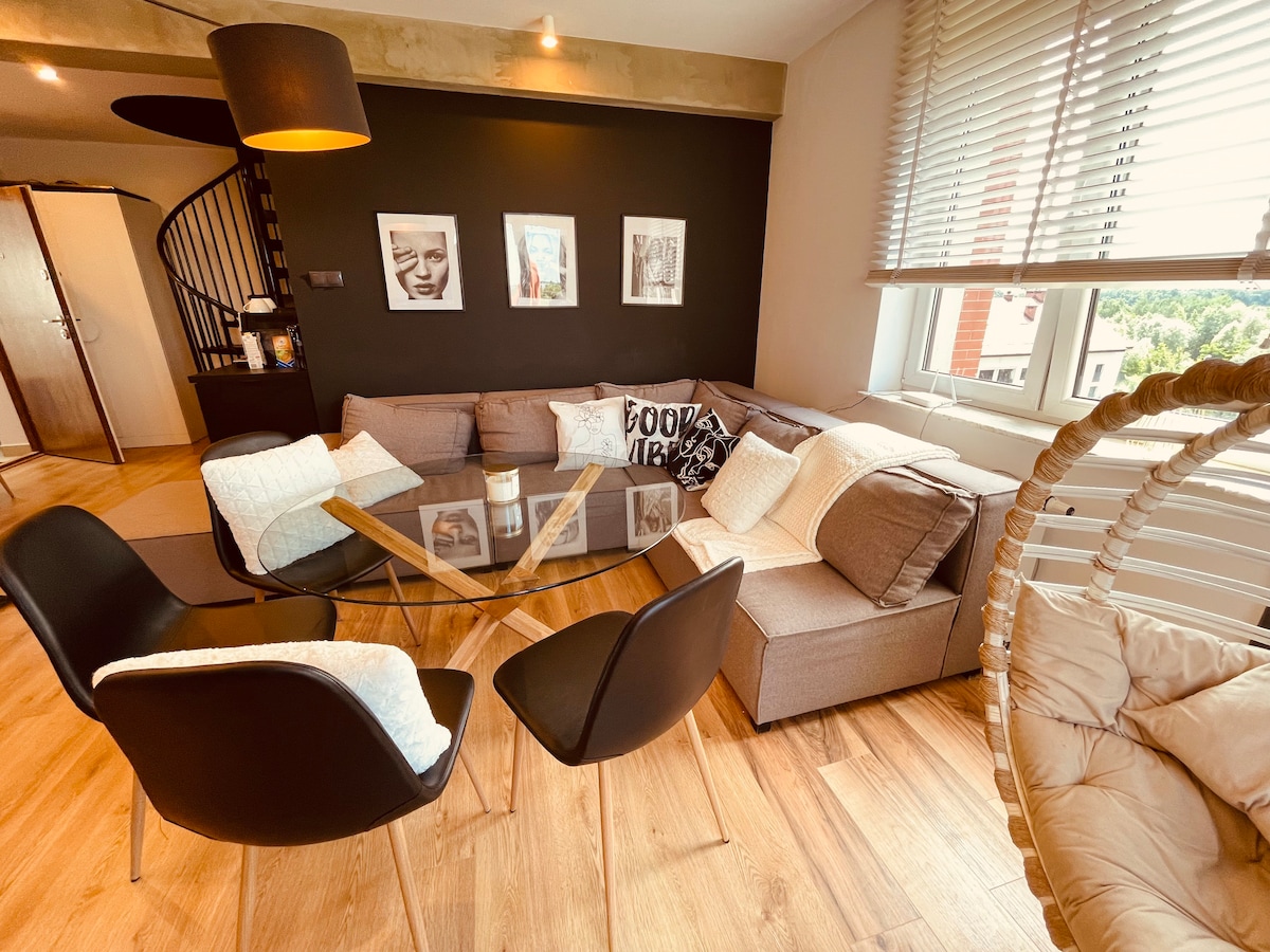 A modern living area features a spacious sectional sofa with neutral-colored cushions, complemented by four stylish black chairs surrounding a glass coffee table. A sleek black wall provides contrast, while natural light enters through large windows adorned with white blinds.