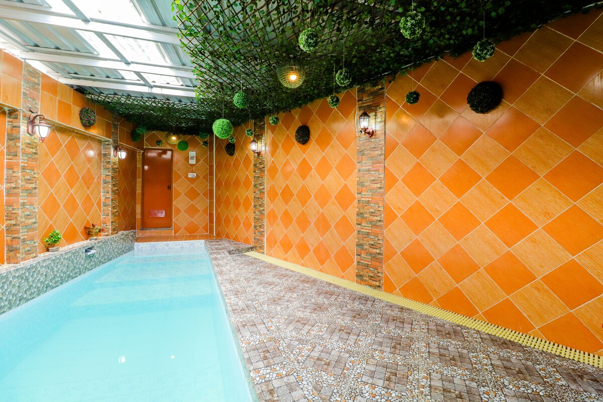 An indoor pool area is surrounded by orange tiled walls, accented with decorative greenery. The turquoise water reflects soft lighting from wall sconces, creating a calm environment. The spacious area features textured flooring, providing a contrasting element to the smooth surface of the pool.