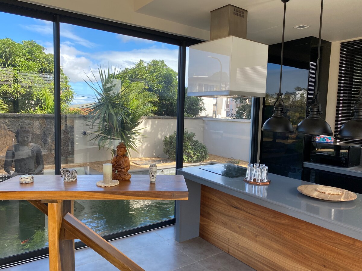 The modern kitchen features a large countertop with warm wooden accents, complemented by stylish pendant lighting. A serene view of the landscaped outdoor area can be seen through expansive glass windows, allowing natural light to fill the space.