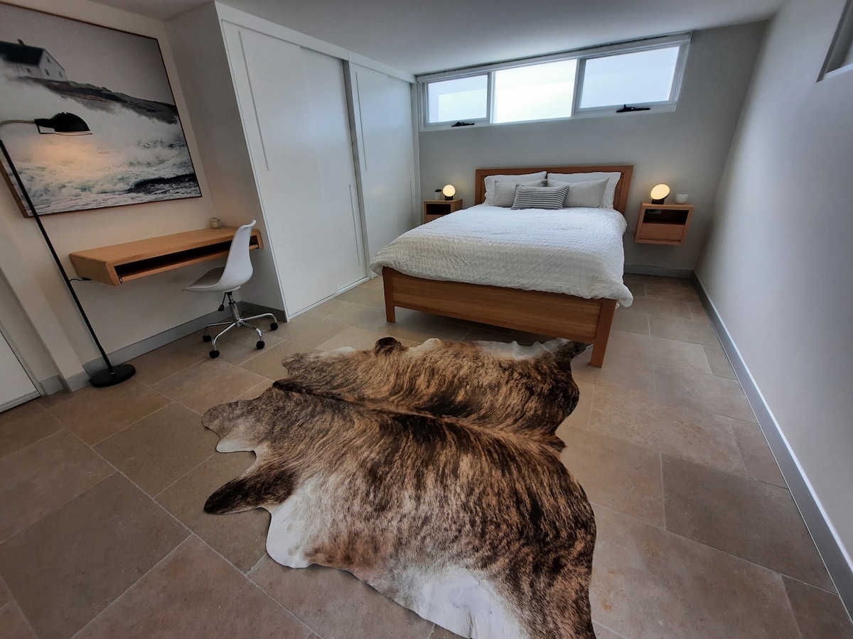 A modern room features a queen bed with a light-colored duvet, flanked by two nightstands with soft lighting. A study area with a sleek desk and chair is present. A large area rug adds warmth to the stone floor, and three windows provide ample natural light.