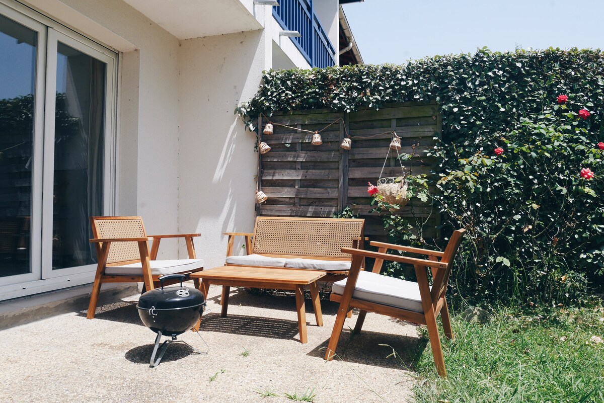 An outdoor seating area features a wooden table surrounded by three chairs with light cushions. A charcoal grill is positioned nearby, and a wooden fence adorned with hanging plants and decorative lights provides a sense of privacy. Lush greenery and blooming flowers enhance the space.