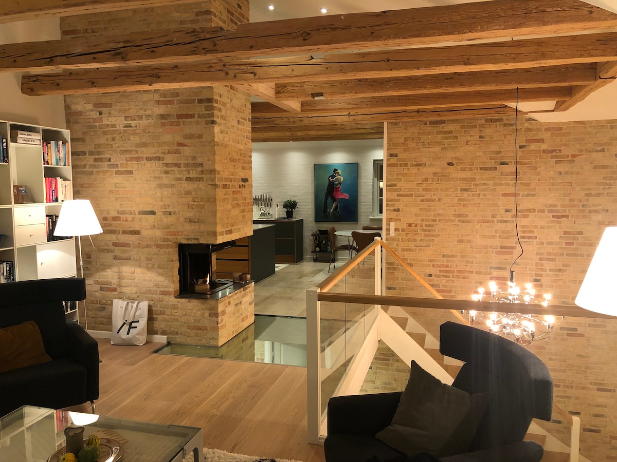 The spacious living area features exposed wooden beams and a warm brick wall, with a modern fireplace enhancing the ambiance. A clear view is provided of the adjoining kitchen, while a stylish staircase leads to the upper level. Soft seating is arranged for comfort.