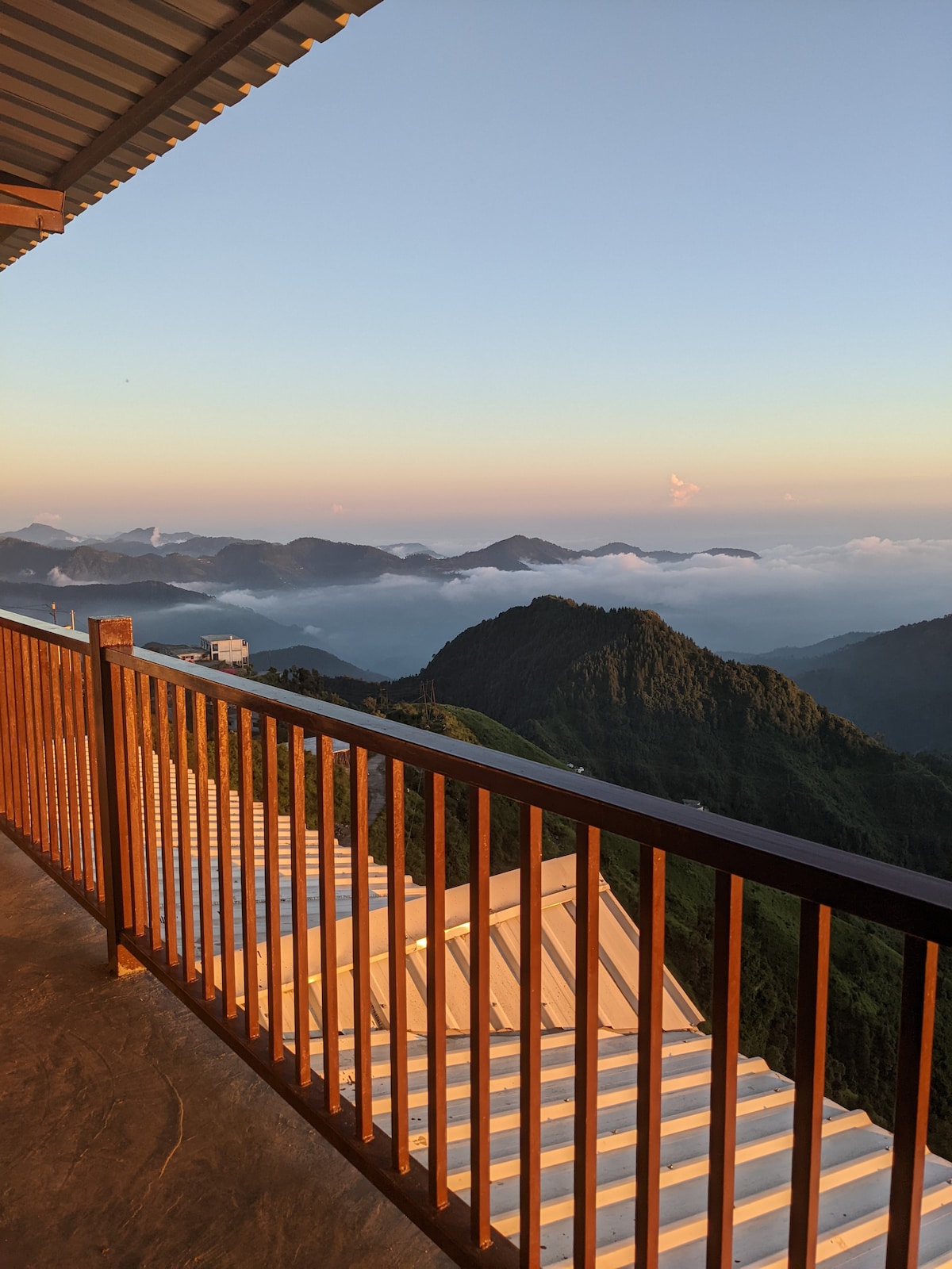 A balcony railing made of warm wood extends over a scenic mountainous landscape. Layers of hills and clouds are visible in the distance, catching the light of the sunset. The roof of the building is partially seen below, adding depth to the view.