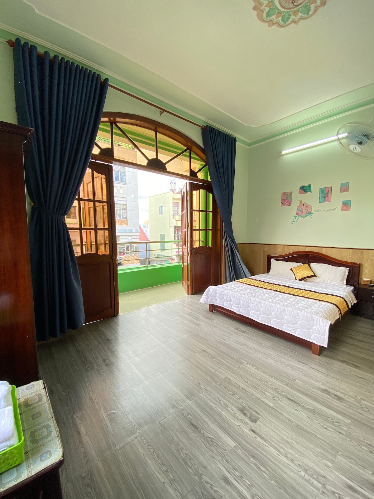 A bright room features a spacious bed with neutral bedding. Large wooden doors open to a small balcony, allowing natural light to fill the space. Dark blue curtains frame the windows, and light wooden flooring complements the room’s design.