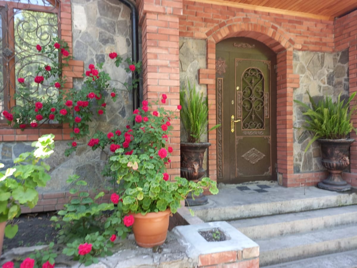 A stone and brick entrance is framed by lush greenery and vibrant red roses. The front door, embellished with decorative metalwork, is flanked by large pots filled with plants. Steps lead up to the doorway, inviting guests inside.