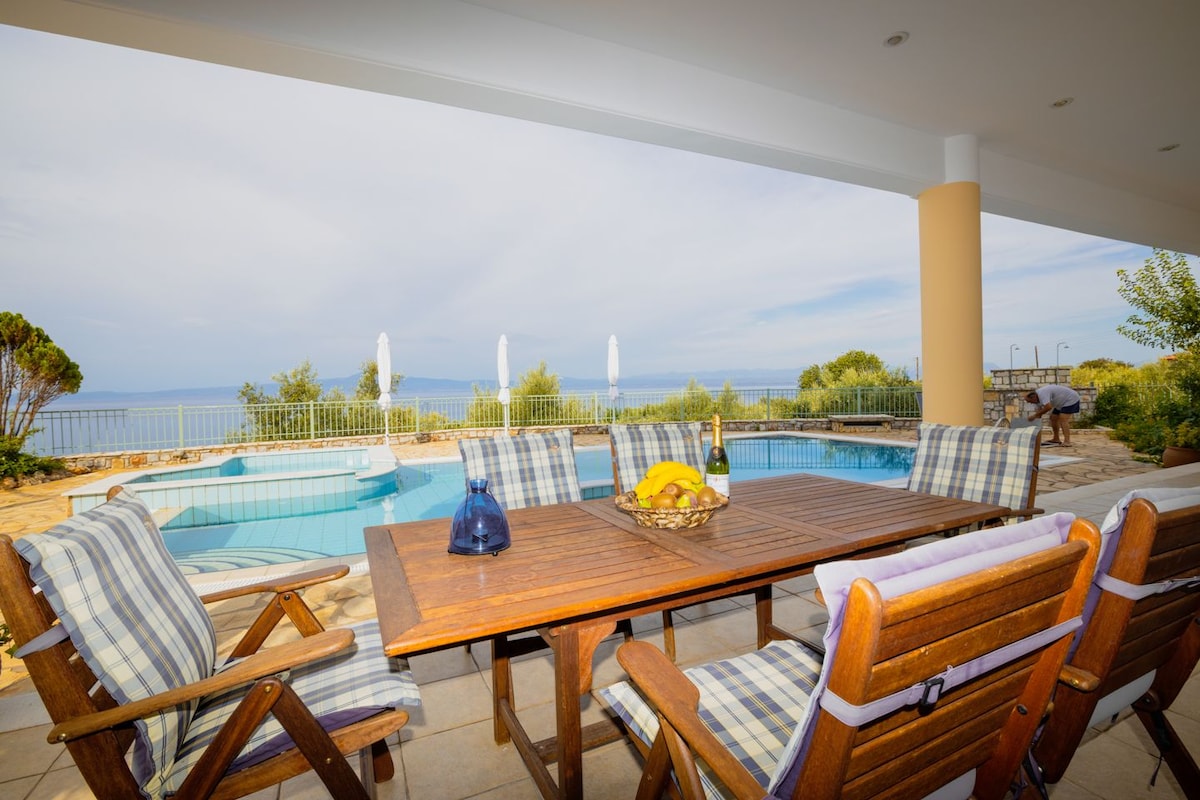 An outdoor dining area features a wooden table surrounded by several chairs, all with striped cushions. A fruit bowl is placed on the table. In the background, a pool is visible, with sun loungers and tall umbrellas under a clear sky.