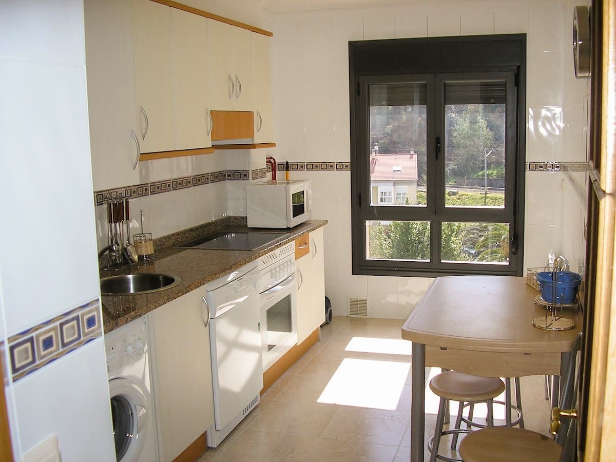 A spacious kitchen is equipped with modern appliances, including a washing machine and microwave. Natural light fills the area through a large window overlooking greenery. A simple wooden table with two stools is positioned near the window, adding a functional space for dining or preparation.