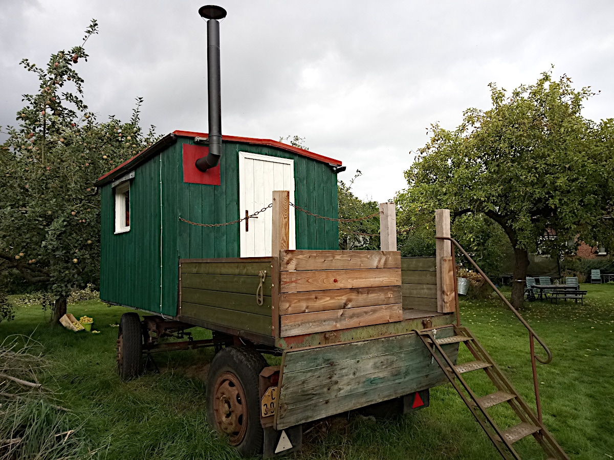 A rustic forest wagon is positioned on a grassy area, featuring a green exterior with a red accent. A chimney extends from the roof, and a set of wooden steps provides access to the platform. Nearby, apple trees and seating areas are visible.