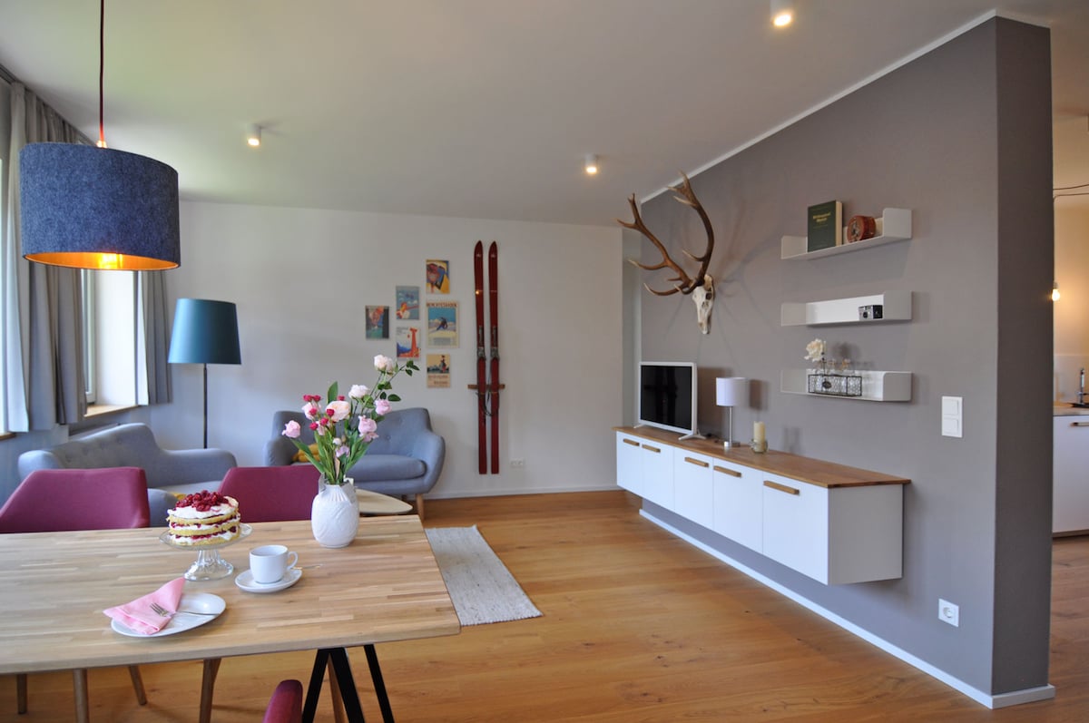 A bright living space features a wooden dining table set for tea with a cake, complemented by a floral centerpiece. Two comfortable seating areas include light blue and pink chairs. Decorative elements, such as vintage skis and a mounted stag's head, add character to the neutral color palette.