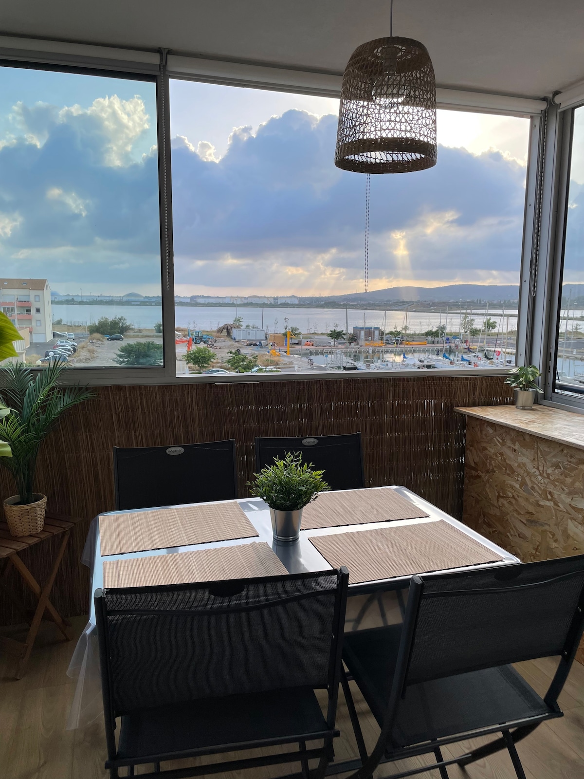 A dining area is positioned near large glass windows, offering views of the waterfront and cloudy sky. A small potted plant decorates the table, which is set with placemats. A woven pendant light hangs above, complementing the natural textures of the space.