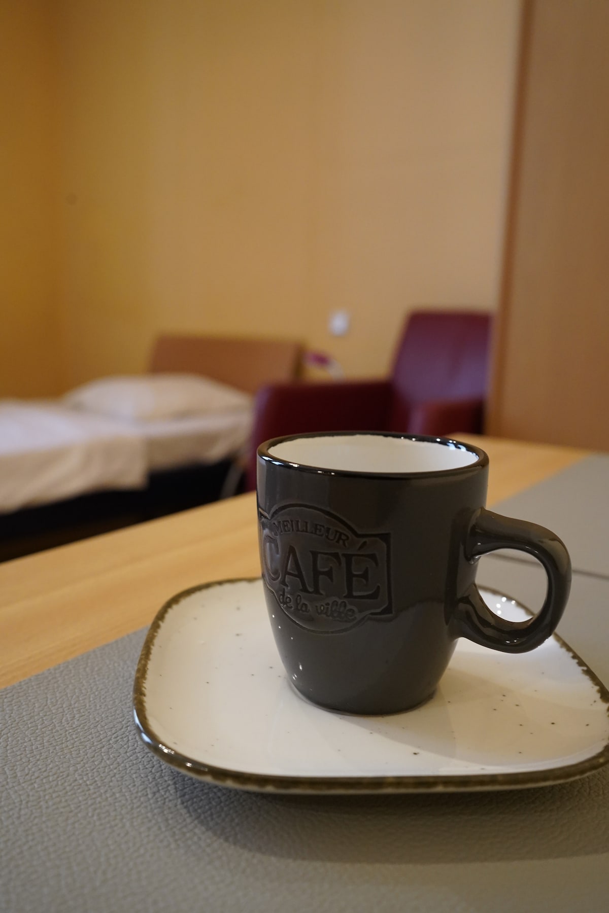 A coffee cup with a decorative handle is placed on a square saucer, resting on a wooden table. In the background, a neutral-colored bedding set is visible on a spacious bed, and two comfortable chairs are positioned nearby.