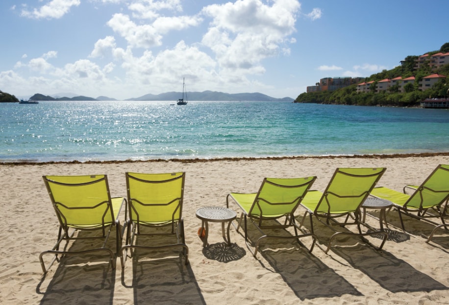 A row of six green beach chairs is arranged on the fine white sand, facing a tranquil shoreline. Gentle waves can be seen lapping against the sand, with clear turquoise waters extending towards distant islands under a bright sky dotted with fluffy clouds.