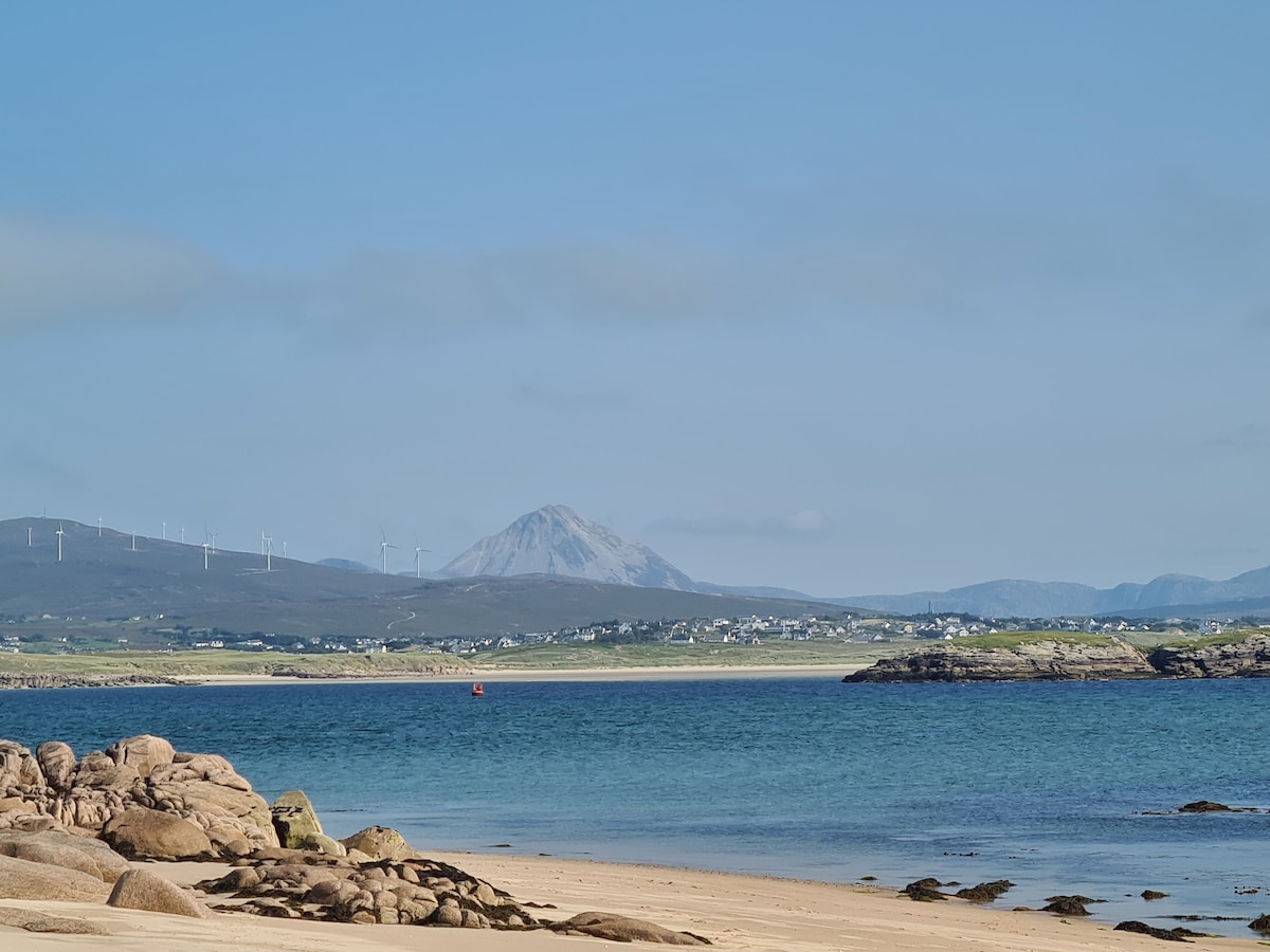 A view of a sandy beach with gentle waves lapping at its shore. Rolling green hills can be seen in the background, along with a mountain peak. Wind turbines dot the landscape, contributing to the serene coastal scenery under a clear blue sky.