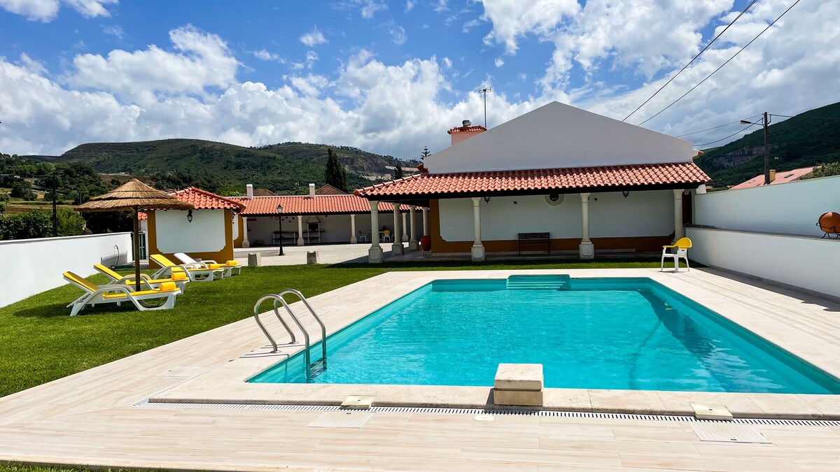 An outdoor area features a clear, inviting swimming pool surrounded by a sun terrace. Yellow loungers are placed on the lawn, and shaded seating areas are available under thatched roofs. A backdrop of rolling hills is visible against a partly cloudy sky.