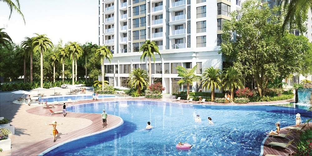 A large outdoor swimming pool is surrounded by palm trees and vibrant landscaping. Sun loungers are positioned around the pool, with various individuals enjoying the water. The modern apartment building looms in the background, creating a welcoming ambiance for relaxation.
