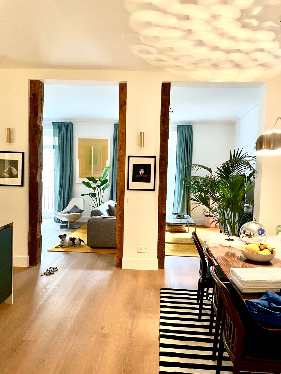 A spacious living area is visible, featuring wooden beams and contemporary decor. A dining table with fruit is placed near the foreground, while a cozy seating area with a sofa and greenery can be seen in the background, illuminated by natural light from large windows.