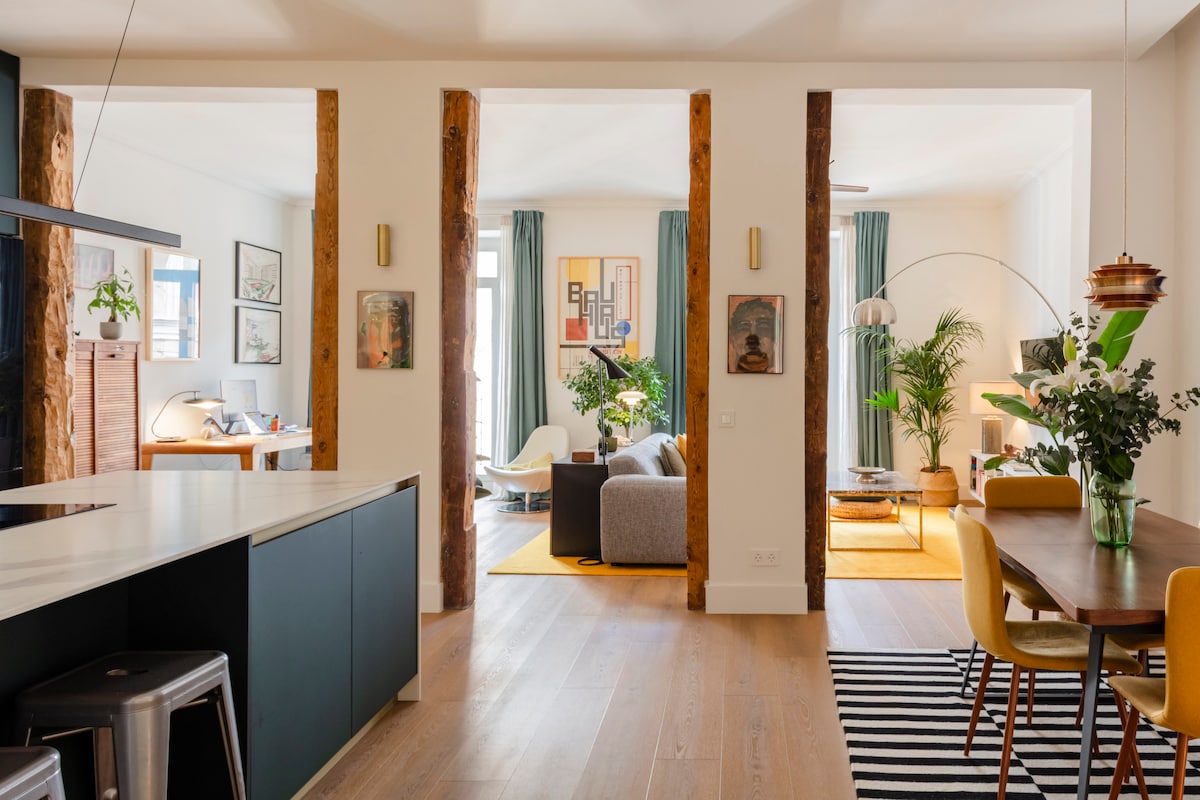 An open living area is divided by wooden beams, creating distinct spaces for dining and relaxation. A modern kitchen with a white countertop is visible, alongside a dining table surrounded by yellow chairs. Soft lighting complements the greenery and artwork throughout the space.