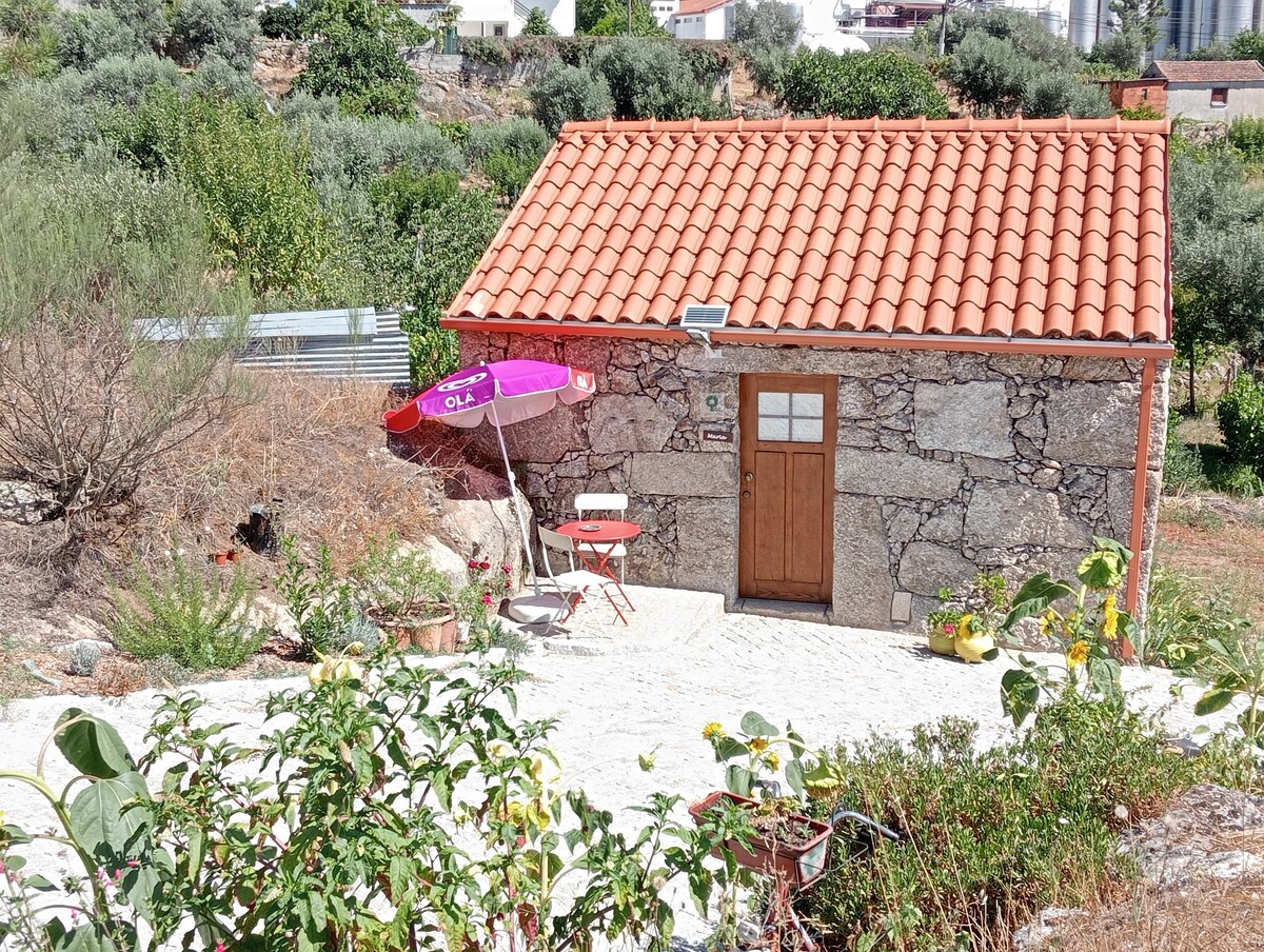 A quaint stone cottage with a red-tiled roof is situated amidst a natural landscape. Outside, a small table and two chairs are positioned under a purple umbrella. Potted plants decorate the area, enhancing the rustic charm of the environment.