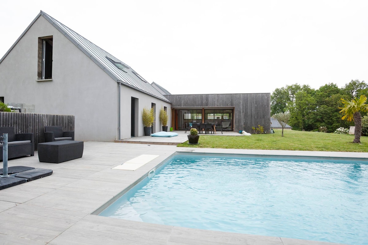 A serene outdoor area features a rectangular pool surrounded by sleek tiles. The house is depicted in the background, showcasing a mix of modern materials. Lush greenery and palm trees enhance the peaceful setting, creating an inviting space for relaxation.