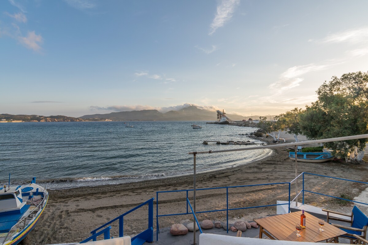 The serene beach view is framed by a wooden railing, showcasing the calm waters and distant mountains. A small boat is visible near the shore, along with several other boats scattered on the water. Sandy beach extends invitingly towards the horizon.