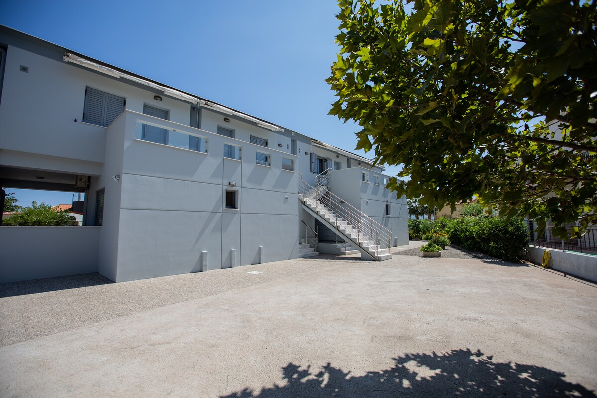 A modern building with a light grey facade is set against a clear blue sky. Stairs lead to upper units, while a spacious outdoor area is surrounded by greenery and landscaping. The ground is composed of gravel, providing a natural pathway to the entrance.