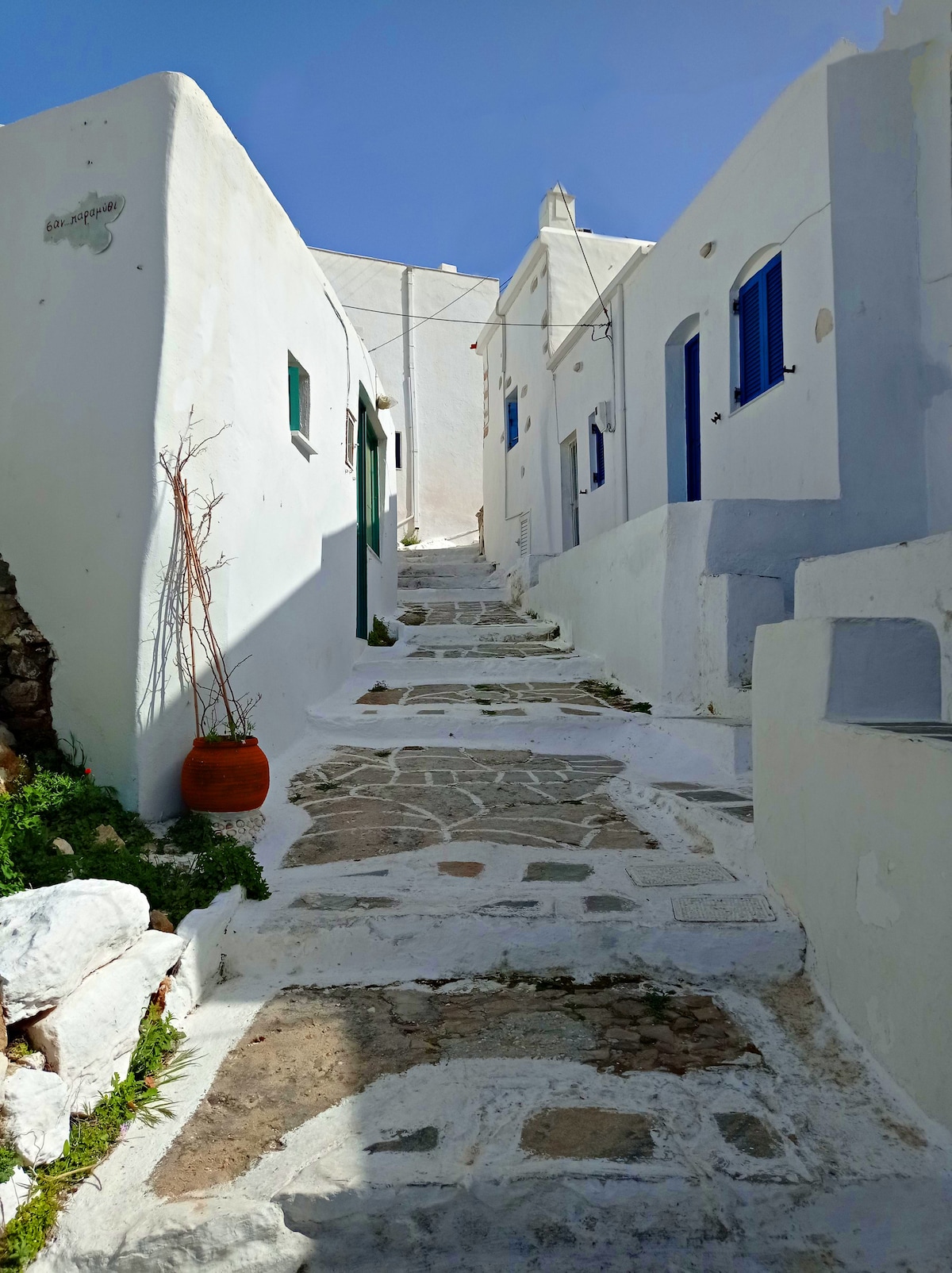 A narrow, stone-paved alley is flanked by whitewashed buildings featuring blue windows and doors. Potted plants add touches of greenery, and shadows are cast against the walls under the sunlight. The path slopes gently upward, leading into the traditional village setting.