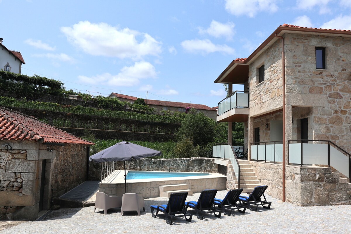 A stone-clad building features a private swimming pool surrounded by lounge chairs. A large umbrella offers shade, while steps lead up to the two-story structure. Greenery is visible in the background, enhancing the outdoor space.
