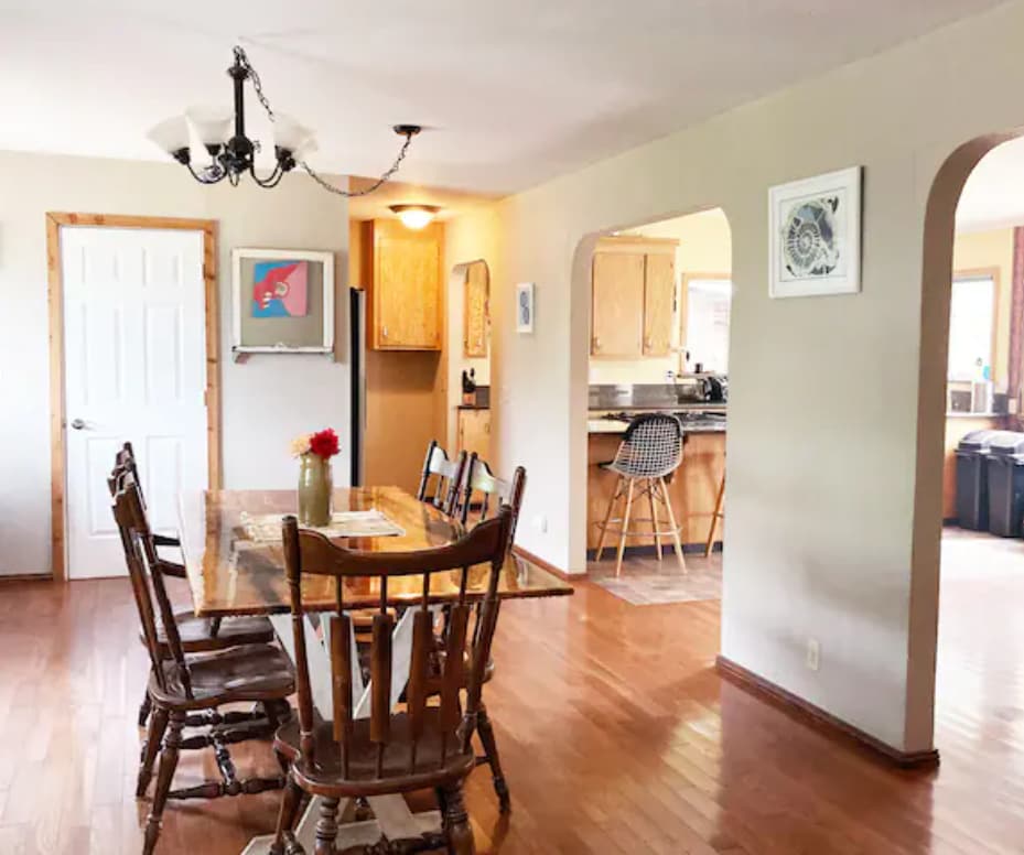 A welcoming dining area features a wooden table surrounded by rustic chairs. Natural light filters through nearby windows, illuminating the space. An open archway leads to a well-equipped kitchen, while contemporary artwork adorns the walls, adding a touch of character to the room.