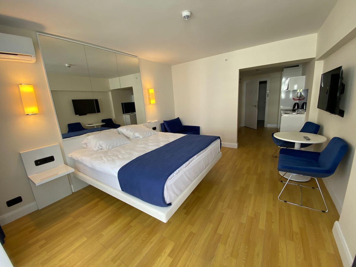 A well-lit room features a floating bed with crisp white linens and a navy blue accent blanket. A mirror wall enhances the spacious feel. A blue armchair and small dining table are positioned nearby, while a television is mounted for entertainment.