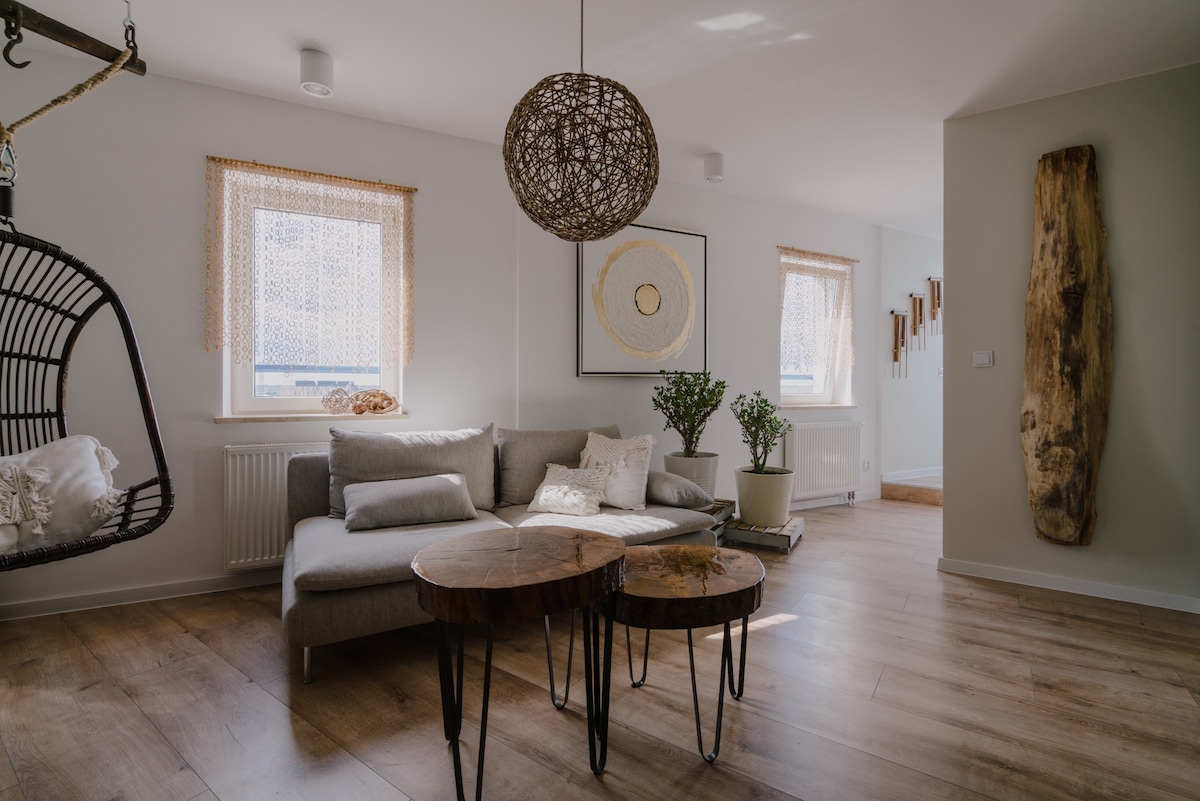 A modern living area features a light gray sectional sofa paired with two round wooden coffee tables. Natural light filters through sheer window coverings, illuminating the space. A decorative plant is placed beside the sofa, and a unique wooden wall art piece is visible on a neutral wall.