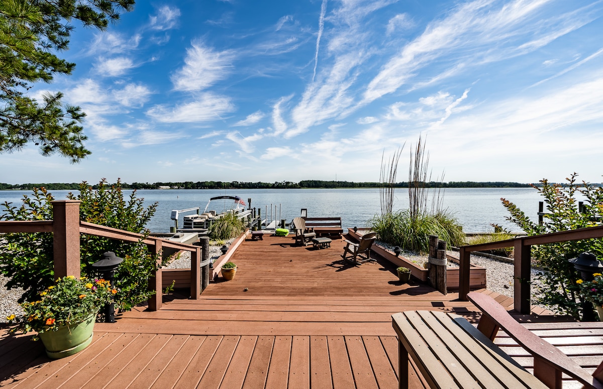 A wooden deck extends toward the calm water, featuring various seating arrangements and potted plants. Lush greenery surrounds the area, while a clear blue sky with wispy clouds enhances the serene setting. A boat dock is visible in the distance.