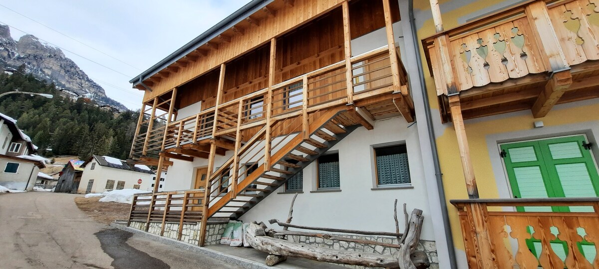 A wooden structure featuring a staircase leading to a second floor, with a balcony adorned with wooden railings. The building's exterior combines stone and painted surfaces, while nearby natural scenery contributes to the charming mountain setting.